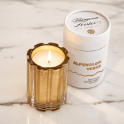 A lit candle in a gold-inlayed fluted glass holder sits beside its white cylindrical packaging container labeled Thompson Ferrier Alpenglow Verge on a white marble surface.