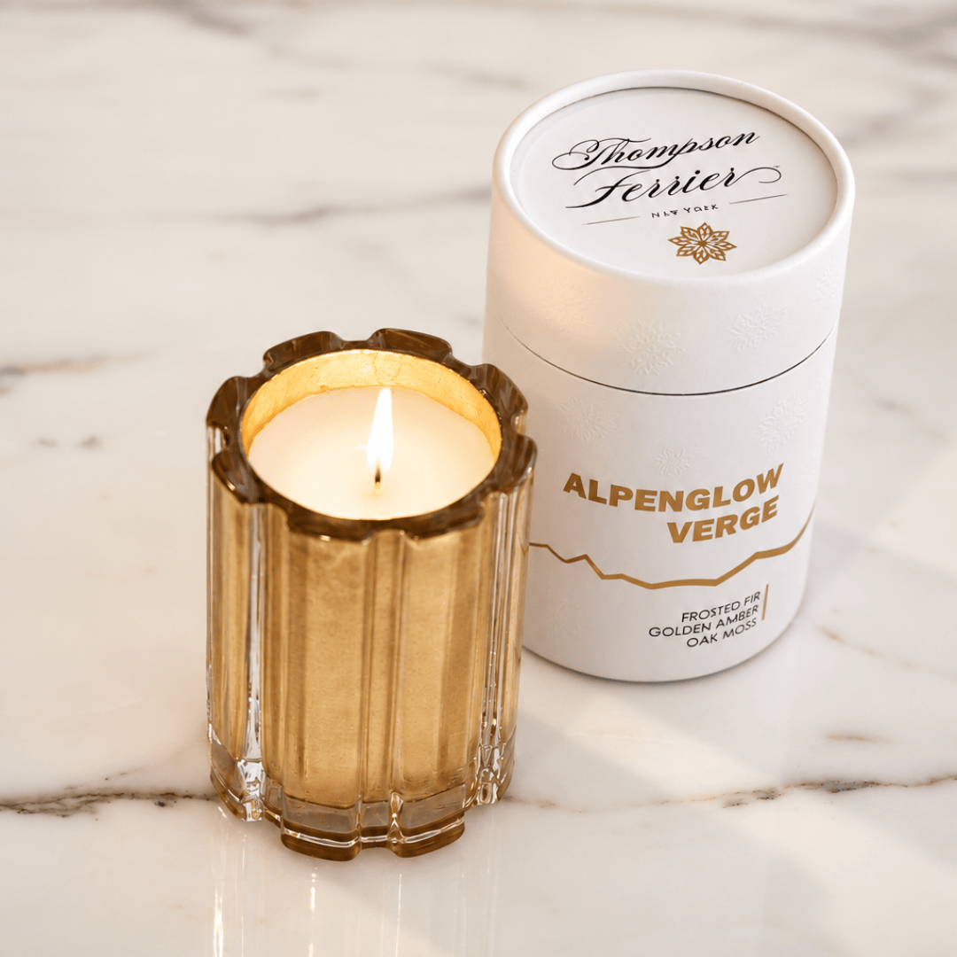 A lit candle in a gold-inlayed fluted glass holder sits beside its white cylindrical packaging container labeled Thompson Ferrier Alpenglow Verge on a white marble surface.
