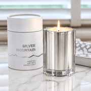 A silver metal candle holder with a lit flame next to its white cylindrical packaging container labeled 'SILVER MOUNTAIN'.