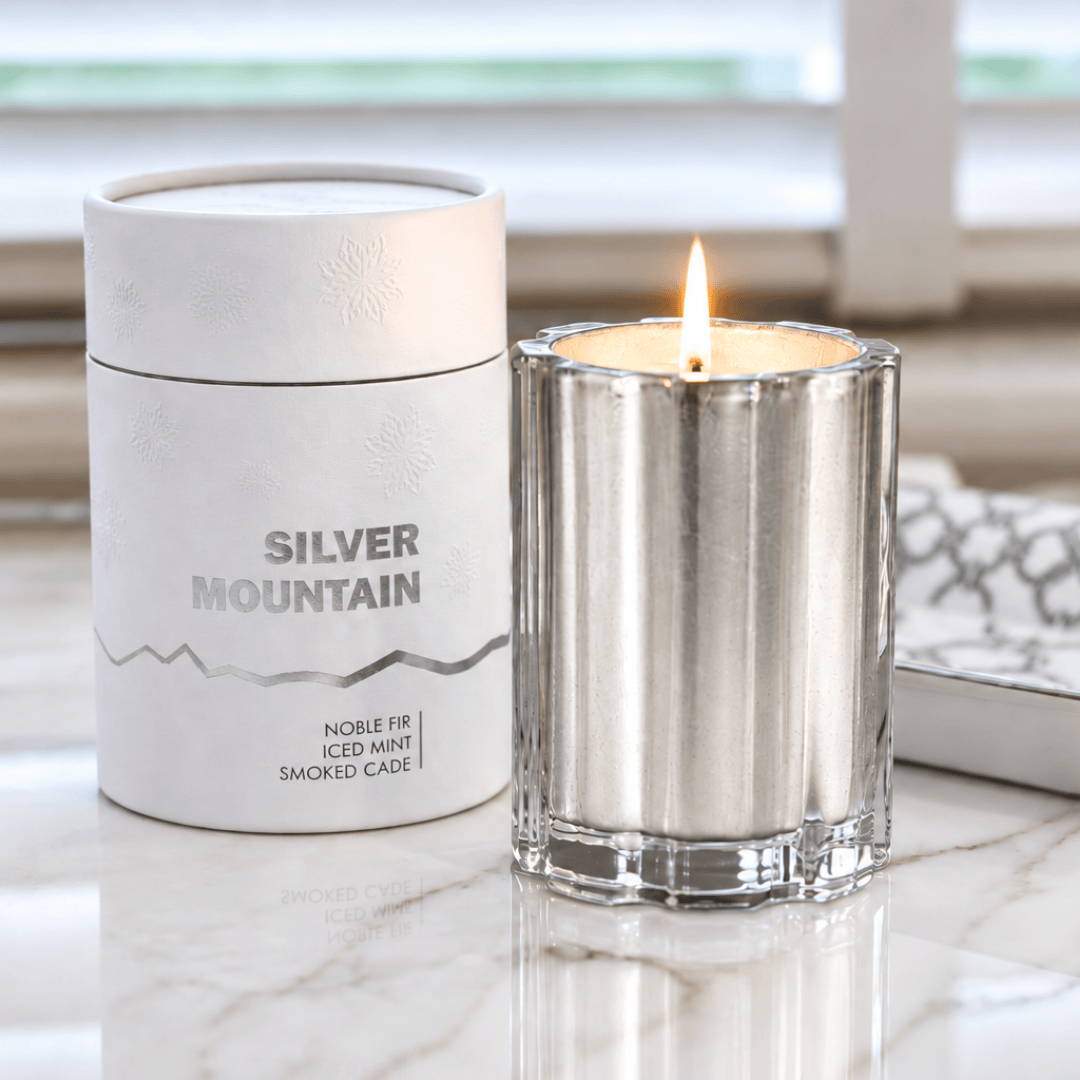A silver metal candle holder with a lit flame next to its white cylindrical packaging container labeled 'SILVER MOUNTAIN'.
