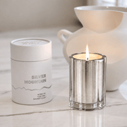 A silver coated glass candle tin labeled 'SILVER MOUNTAIN' sits beside a lit silver candle holder with a small flame, on a light marble surface with a white ceramic pitcher blurred in the background.
