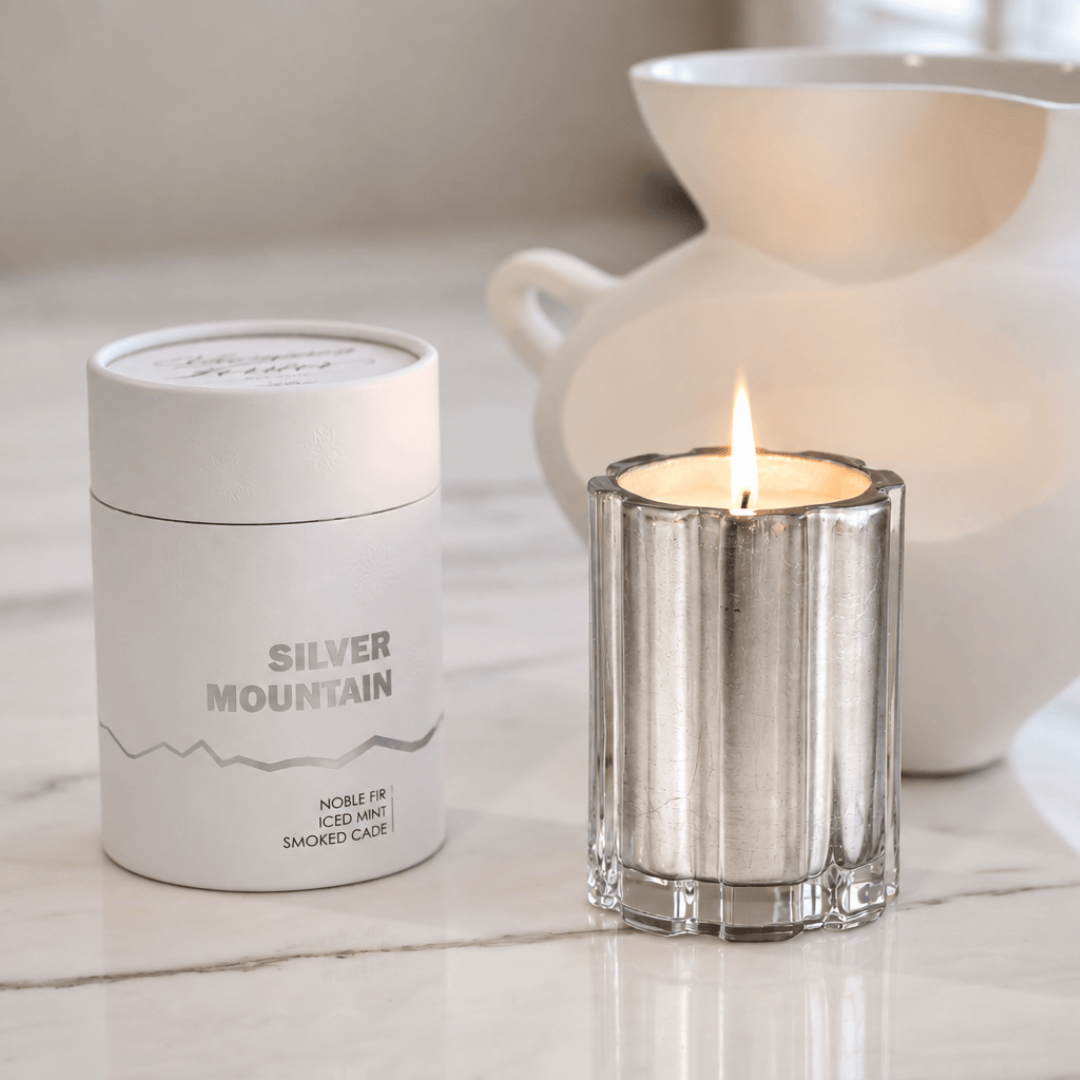 A silver coated glass candle tin labeled 'SILVER MOUNTAIN' sits beside a lit silver candle holder with a small flame, on a light marble surface with a white ceramic pitcher blurred in the background.