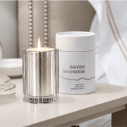 A lit silver glass candle in a clear glass holder sits beside a white cylindrical tin labeled 'SILVER MOUNTAIN' on a light tabletop in a softly styled interior setting.