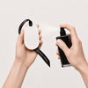 Two hands holding a white oval-shaped diffuser with a black ribbon and a black spray bottle against a light beige background.