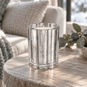 A clear ribbed glass candle holder with a textured, metallic silver finish, containing a white candle, set on a rustic wooden table in a cozy living room setting.